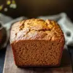 Pumpkin bread gf - the image shows a freshly baked loaf of bread on a wooden cutting board. the bread appears to be golden brown in color and has a crumbly texture. it is sitting on a dark wooden table with a white cloth napkin and a small pumpkin in the background. the overall mood of the image is rustic and cozy.