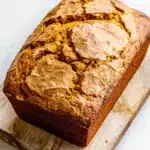 Pumpkin bread high altitude - the image shows a freshly baked loaf of bread on a wooden cutting board. the bread appears to be golden brown in color and has a crumbly texture on top. the slices of bread are arranged in a circular pattern, with some overlapping each other. the cutting board is placed on a white marble countertop. the background is blurred, making the bread the focal point of the image.