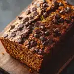 Pumpkin zucchini chocolate bread - the image shows a freshly baked loaf of bread on a wooden cutting board. the bread appears to be a dark brown color and has a smooth texture. it is topped with chocolate chips scattered across the top, giving it a shiny appearance. the loaf is cut in half, revealing the moist and fluffy interior. the cutting board is placed on a dark grey countertop.