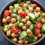 Quinoa and chickpea salad - the image is a close-up of a bowl of salad. the bowl is made of dark brown ceramic and is placed on a dark grey surface. the salad is made up of small, round chickpeas, red onions, sliced cucumbers, and cherry tomatoes. there are also some green herbs scattered throughout the salad, adding a pop of color to the dish. the overall appearance is fresh and appetizing.