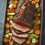 Roast beef marinade slow cooker - the image shows a rectangular baking dish filled with a roast beef dish. the roast beef is cooked medium-rare and has a golden brown crust on top. it is garnished with sliced carrots, celery, and onions. the dish is served on a dark grey countertop. the vegetables are arranged in a way that they are overlapping each other, creating a colorful and appetizing presentation.
