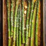 Roasted asparagus garlic - the image shows a baking tray with cooked asparagus spears on it. the tray is rectangular in shape and is placed on a wooden table. the spears are arranged in a single layer on the tray, with some of them overlapping each other. they are covered in a light green sauce and sprinkled with parmesan cheese. there are also some small pieces of garlic scattered around the tray. the overall appearance of the dish is fresh and appetizing.