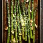 Roasted asparagus garlic - the image shows a baking tray with cooked asparagus spears on it. the spears are arranged in a single layer on the tray, with some of them overlapping each other. the tray is lined with parchment paper and there are small pieces of garlic scattered around the spears. the garlic appears to be seasoned with herbs and spices, giving the spears a golden brown color. the background is a dark wooden surface, possibly a table or countertop.