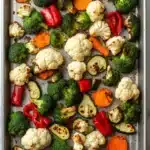 Roasted vegetables in ninja air fryer - the image shows a baking tray filled with roasted vegetables. the tray is lined with parchment paper and is placed on a white marble countertop. the vegetables are arranged in a single layer on the tray, with some overlapping each other. there are broccoli, cauliflower, carrots, zucchini, and red bell peppers scattered throughout the tray. the colors of the vegetables are vibrant and the tray appears to be freshly baked.