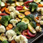 Roasted vegetables in ninja air fryer - the image shows a tray of roasted vegetables on a wooden table. the vegetables are arranged in a single layer on the tray, with some overlapping each other. the tray appears to be made of metal and has a dark brown color. the vegetables include broccoli, cauliflower, red and yellow bell peppers, zucchini, and carrots. the broccoli is cut into small pieces and the bell peppers are cut into thin slices. the carrots are bright orange and appear to be seasoned with herbs and spices. the cauliflower is white and fluffy, while the peppers are a mix of red, yellow, and green colors. the red bell pepper is slightly larger than the yellow bell pepper and is slightly smaller than the green bell pepper. the other vegetables are also roasted and have a golden brown color, indicating that they have been roasted to perfection.