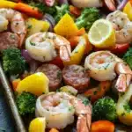 Sausage and shrimp sheet pan meal - the image shows a tray of cooked shrimp and vegetables. the tray is lined with parchment paper and is placed on a black countertop. the shrimp are pink and appear to be seasoned with herbs and spices. the vegetables include broccoli, red onions, yellow bell peppers, and red bell peppers. there are also slices of lemon scattered throughout the dish. the dish looks appetizing and ready to eat.