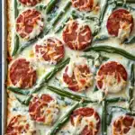 Sausage green beans alfredo sheet pan - the image shows a rectangular baking dish filled with a dish of green beans and pepperoni. the dish is covered in a creamy white sauce and topped with melted cheese. the green beans are long and slender, and they are arranged in a single layer on top of the sauce. the pepperoni is round and red, and it appears to be seasoned with herbs and spices. the cheese is melted and bubbly, and there are small pieces of pepperoni scattered throughout the dish. the baking dish is sitting on a black countertop.