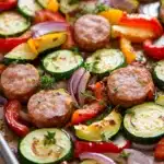 Sausage peppers and zucchini sheet pan - the image shows a tray of grilled vegetables on a wooden table. the vegetables appear to be zucchini, red and yellow bell peppers, and red onions. there are also small pieces of sausage scattered throughout the tray. the sausage is golden brown and looks crispy on the outside. the tray is lined with parchment paper and there is a sprig of thyme on top. the background is blurred, but it appears to be a kitchen countertop.