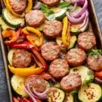 Sausage peppers and zucchini sheet pan - the image shows a rectangular baking tray filled with grilled meatballs and vegetables. the meatballs are golden brown and appear to be seasoned with herbs and spices. the vegetables include zucchini, red onions, yellow bell peppers, and red onions. the tray is garnished with fresh basil leaves. the background is a dark grey countertop.