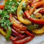 Sauteed fajita vegetables - the image is a close-up of a plate of food. the plate is white with a speckled pattern and is placed on a wooden table. the food appears to be a stir-fry dish with sliced red and yellow bell peppers, onions, and cilantro. the peppers are arranged in a circular pattern and are garnished with fresh cilantro leaves. the colors of the peppers are vibrant and the dish looks fresh and appetizing.