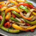 Sauteed fajita vegetables - the image is a close-up of a plate of stir-fry vegetables. the vegetables are sliced and arranged in a circular pattern on a dark grey plate. the colors of the vegetables include red, yellow, green, and orange bell peppers, as well as sliced onions. the plate is sitting on a wooden table with a rustic background. the dish appears to be freshly cooked and ready to eat.