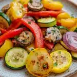 Sauteed vegetables in oven - the image is a close-up of a plate of colorful vegetables. the plate is white with a speckled pattern and is placed on a wooden table. the vegetables are arranged in a circular pattern on the plate. on top of the vegetables, there are sliced red and yellow bell peppers, zucchini, and mushrooms. the bell peppers are cut into thin slices and are garnished with chopped herbs. the mushrooms are dark brown and appear to be seasoned with herbs and spices. there are also slices of red onion scattered throughout the dish. the overall appearance of the dish is fresh and appetizing.