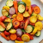 Sauteed vegetables in oven - the image is a close-up of a colorful salad in a white bowl with a speckled pattern. the salad is made up of various types of vegetables, including zucchini, red onions, yellow squash, and red bell peppers. the vegetables are arranged in a circular pattern on the bowl, with some overlapping each other. the colors of the vegetables are vibrant and the dish looks fresh and appetizing. the bowl is sitting on a wooden table.