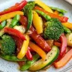 Sauteed vegetables in sauce - the image is a close-up of a plate of stir-fry vegetables. the plate is white with a speckled pattern and is placed on a wooden table. the vegetables are mixed together in a colorful and appetizing manner. the colors of the vegetables include red, yellow, green, and orange bell peppers, as well as sliced zucchini and broccoli. there are also some sesame seeds sprinkled on top. the dish appears to be freshly cooked and ready to eat.