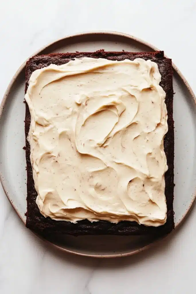 Sheet cake with cream cheese frosting - the image shows a square brownie on a white marble plate. the brownie appears to be freshly baked and has a rich, dark chocolate color. on top of the brownie, there is a generous layer of creamy white frosting that is spread evenly across the top. the frosting has a smooth texture and looks smooth and shiny. the plate is round and has an oval shape.