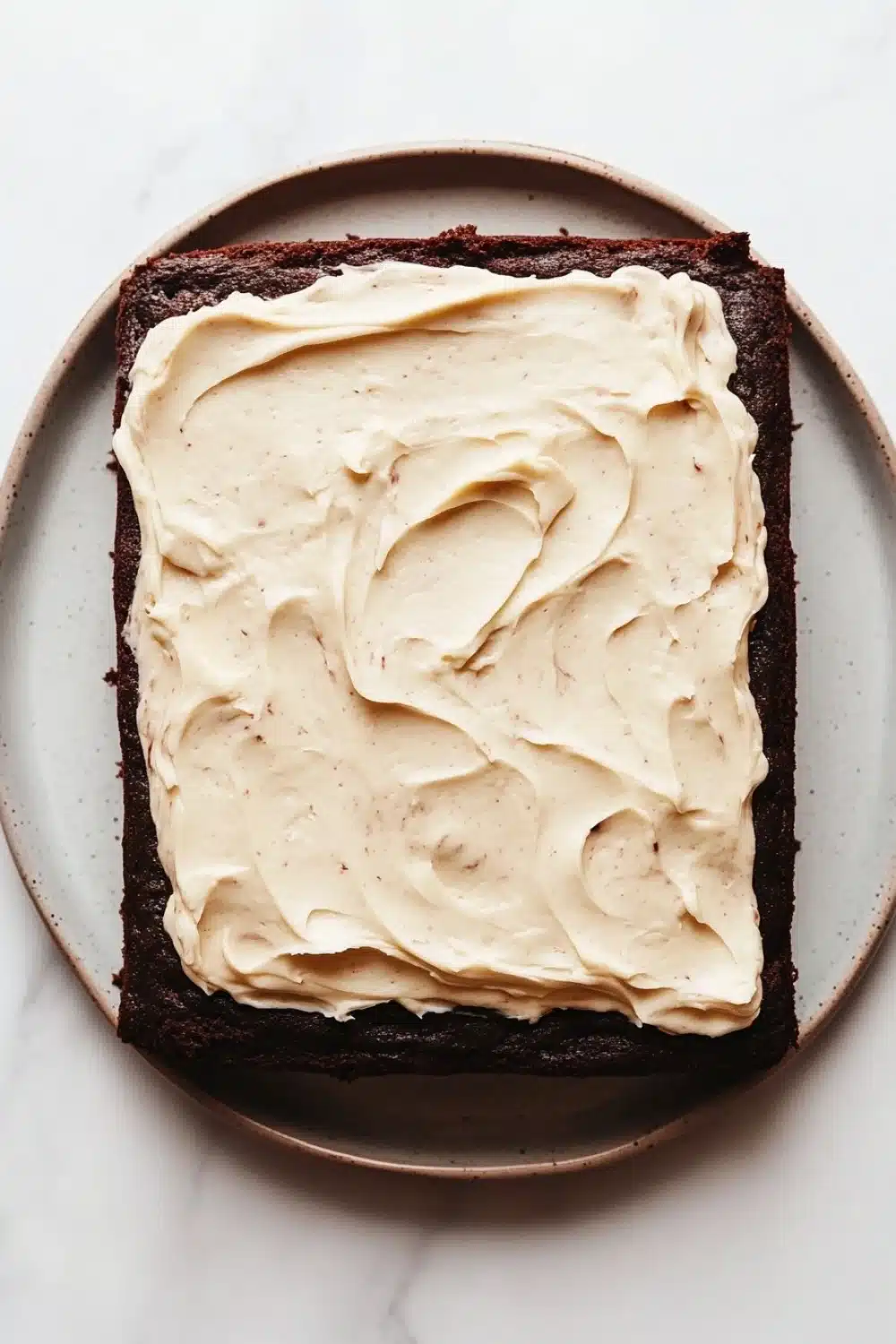 Sheet cake with cream cheese frosting - the image shows a square brownie on a white marble plate. the brownie appears to be freshly baked and has a rich, dark chocolate color. on top of the brownie, there is a generous layer of creamy white frosting that is spread evenly across the top. the frosting has a smooth texture and looks smooth and shiny. the plate is round and has an oval shape.