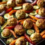 Sheet pan chicken meatballs and vegetables - the image shows a tray of roasted vegetables on a wooden table. the vegetables are arranged in a circular pattern on the tray, with some overlapping each other. the meatballs are golden brown and appear to be seasoned with herbs and spices. they are cut into small cubes and are arranged on top of a bed of sliced cucumbers, red onions, and red bell peppers. the tray is lined with parchment paper and there is a sprig of rosemary on the side. the background is blurred, but it appears to be a kitchen countertop.