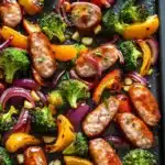 Sheet pan chicken sausage honey garlic - the image shows a tray of cooked sausages and vegetables. the tray is lined with black parchment paper and is filled with a variety of colorful vegetables, including broccoli, red onions, yellow bell peppers, and red onions. the vegetables are arranged in a circular pattern on the tray, with some overlapping each other. the sausage is golden brown and appears to be seasoned with herbs and spices. the colors of the vegetables are vibrant and the dish looks appetizing.