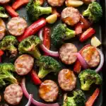 Sheet pan chicken sausage honey garlic - the image shows a black baking tray filled with various types of food. on the tray, there are several pieces of sausage, broccoli, red and yellow bell peppers, and red onions. the sausage appears to be cooked and has a golden brown color. the vegetables are arranged in a circular pattern around the sausage and broccoli. the tray is sitting on a black countertop.