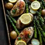 Sheet pan dinner with asparagus - the image shows a baking tray filled with grilled chicken and asparagus. the chicken is golden brown and appears to be seasoned with herbs and spices. the asparaglips are bright green and look fresh and vibrant. there are also small potatoes scattered throughout the tray, adding a pop of color to the dish. the tray is lined with parchment paper and is placed on a dark wooden table. the overall presentation of the dish is appetizing and visually appealing.