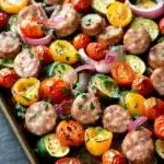 Sheet pan dinners with chicken sausage - the image shows a tray of grilled meatballs and vegetables. the meatballs are arranged in a circular pattern on the tray, with some overlapping each other. the vegetables include sliced tomatoes, zucchini, and bell peppers. the tomatoes are bright red and appear to be seasoned with herbs and spices. the bell peppers are yellow and have a slightly charred texture. the tray is lined with parchment paper, and the background is dark, making the colors of the vegetables stand out.