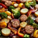 Sheet pan dinners with chicken sausage - the image shows a tray of cooked vegetables on a white countertop. the vegetables appear to be roasted potatoes, broccoli, red and yellow bell peppers, and sausage. the potatoes are golden brown and have a crispy texture. the broccoli is bright green and the bell peppers are red. the sausage is browned and appears to be seasoned with herbs and spices. the tray is lined with parchment paper and is filled to the brim with the vegetables.