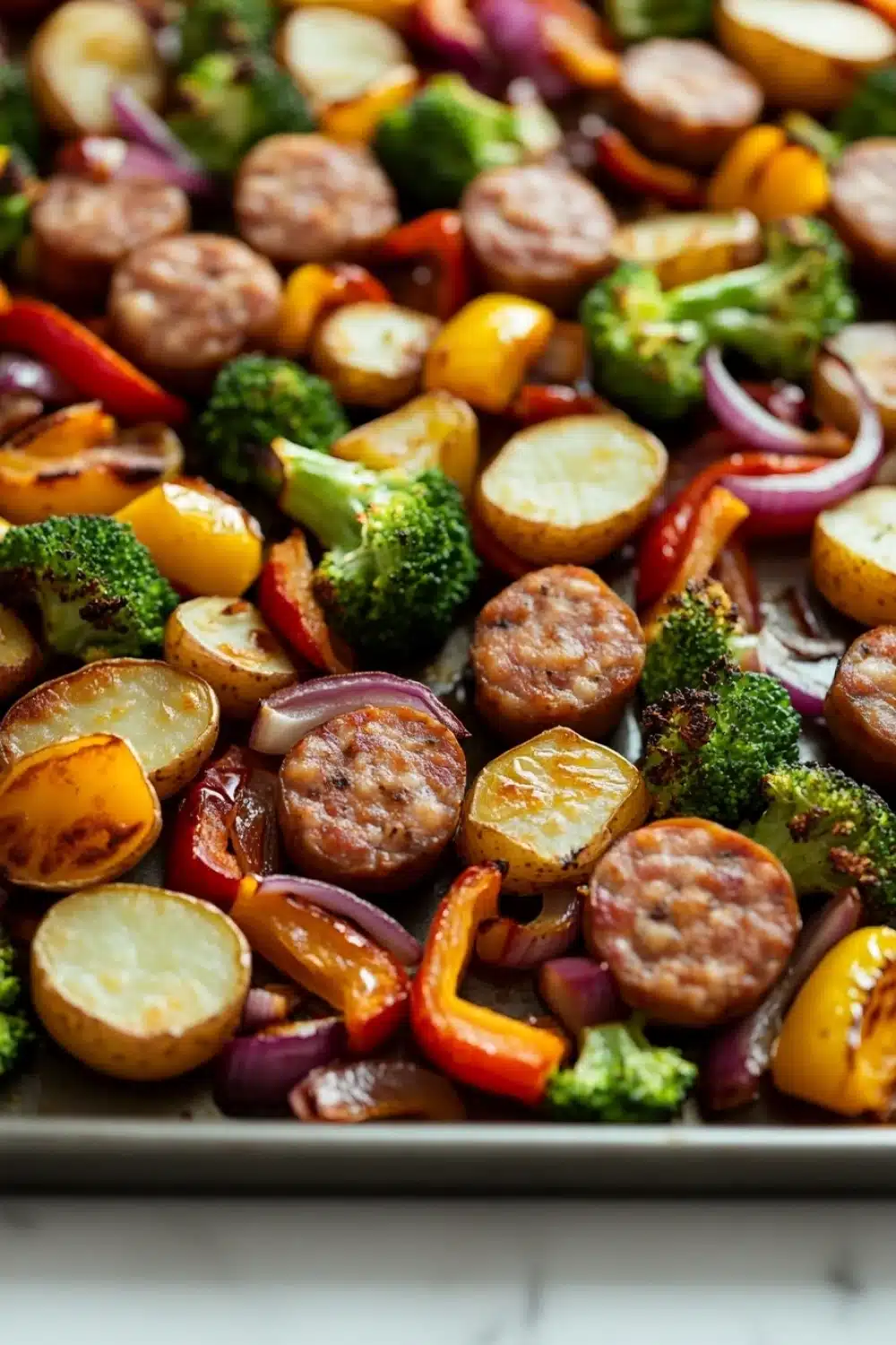 Sheet pan dinners with chicken sausage - the image shows a tray of cooked vegetables on a white countertop. the vegetables appear to be roasted potatoes, broccoli, red and yellow bell peppers, and sausage. the potatoes are golden brown and have a crispy texture. the broccoli is bright green and the bell peppers are red. the sausage is browned and appears to be seasoned with herbs and spices. the tray is lined with parchment paper and is filled to the brim with the vegetables.