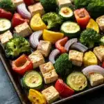 Sheet pan tofu and vegetables - the image shows a rectangular baking tray filled with various types of vegetables and tofu. the vegetables include broccoli, zucchini, bell peppers, and red onions. the tofu is cut into cubes and appears to be seasoned with herbs and spices. the tray is lined with parchment paper and is placed on a dark grey countertop. the overall appearance of the dish is colorful and appetizing.