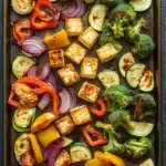 Sheet pan tofu and vegetables - the image shows a baking tray filled with various types of vegetables and tofu. the tray is lined with parchment paper and is placed on a dark grey countertop. on the left side of the tray, there are sliced zucchini, red onions, and yellow bell peppers, while on the right side, there is a pile of broccoli florets. the vegetables are arranged in a circular pattern, with some overlapping each other. the tofu is cubes of tofu and appears to be seasoned with herbs and spices. the colors of the vegetables are vibrant and the tray looks freshly cooked.