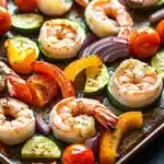 Sheet pan vegetables and shrimp - the image shows a tray of cooked shrimp and vegetables. the shrimp are arranged in a circular pattern on the tray, with some overlapping each other. the vegetables include sliced zucchini, cherry tomatoes, red onions, and yellow bell peppers. the colors of the vegetables are vibrant and the dish appears to be seasoned with herbs and spices. the tray is sitting on a dark blue countertop.