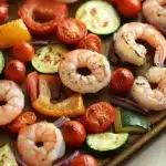 Sheet pan vegetables and shrimp - the image shows a tray of cooked shrimp and vegetables. the shrimp are arranged in a circular pattern on the tray, with some overlapping each other. the vegetables include sliced zucchini, cherry tomatoes, red onions, and yellow bell peppers. the colors of the vegetables are vibrant and the shrimp appear to be seasoned with herbs and spices. the tray is lined with parchment paper, and the background is a wooden table.