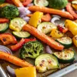 Sheet pan vegetables with carrots - the image shows a baking tray filled with roasted vegetables. the vegetables are arranged in a circular pattern on the tray, with some overlapping each other. the carrots are orange and appear to be seasoned with herbs and spices. there are also small pieces of red and yellow bell peppers, broccoli, and zucchini scattered throughout the tray. the tray is sitting on a wooden table, and the background is blurred, but it appears to be a kitchen countertop.
