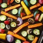 Sheet pan vegetables with carrots - the image shows a baking tray filled with roasted vegetables. the vegetables are arranged in a circular pattern on the tray, with some overlapping each other. the carrots are orange and appear to be seasoned with herbs and spices. there are also sliced red onions, yellow bell peppers, and mushrooms scattered throughout the tray. the tray is lined with parchment paper, and the vegetables are glistening with oil. the background is dark, making the vegetables stand out.