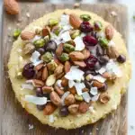 Shortbread cookie toppings - the image shows a round cake on a wooden cutting board. the cake appears to be a light yellow color and has a crumbly texture. on top of the cake, there are various nuts scattered around, including almonds, pistachios, and cranberries. the nuts are arranged in a scattered manner, with some overlapping each other. the cutting board is placed on a white marble countertop.