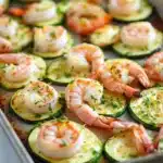 Shrimp and zucchini sheet pan dinner - the image shows a baking tray lined with zucchini boats. the zucchinis are arranged in a single layer on the tray, with some of them overlapping each other. the shrimp are pink and appear to be seasoned with herbs and spices. the tray is sitting on a white marble countertop.