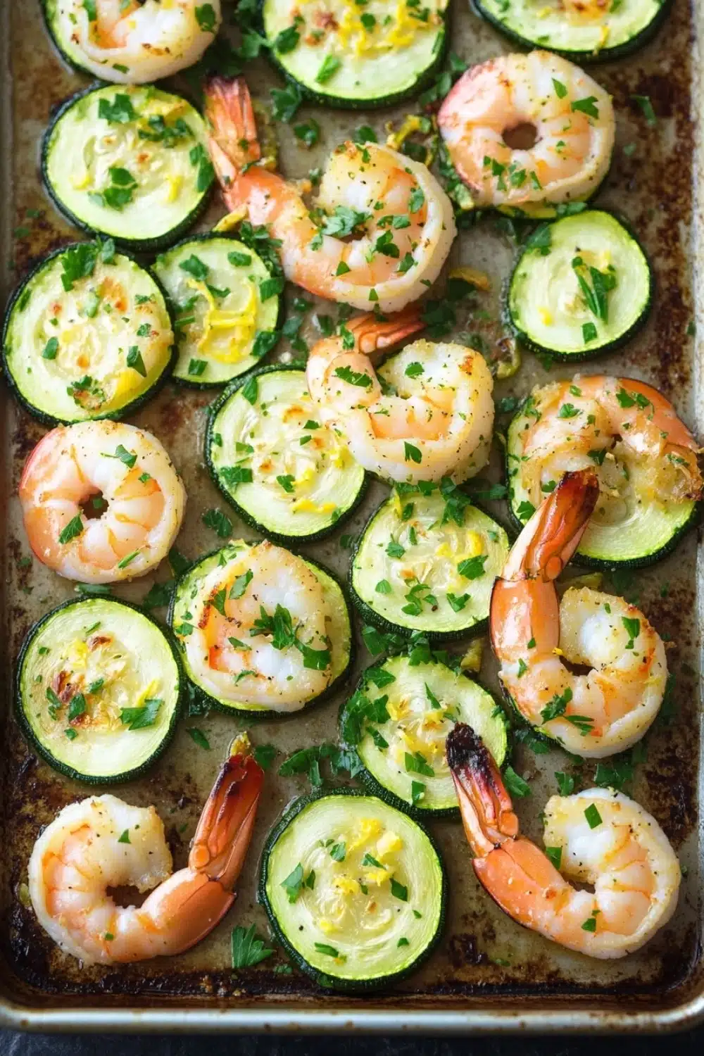 Shrimp and zucchini sheet pan dinner - the image shows a baking tray filled with cooked shrimp and zucchini. the shrimp are pink and appear to be seasoned with herbs and spices. the zucchinis are sliced and arranged in a circular pattern on the tray. the tray is lined with parchment paper and there are a few sprigs of parsley scattered around the tray, adding a pop of green color to the dish. the background is dark, making the colors of the dish stand out.