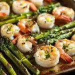 Shrimp sheet pan with asparagus - the image shows a baking tray with cooked shrimp and asparagus. the shrimp are arranged in a single layer on the tray, with some of them overlapping each other. the asparaglips are bright green and appear to be seasoned with herbs and spices. the tray is sitting on a wooden table, and there is a white plate in the background. the overall presentation of the dish is appetizing and visually appealing.