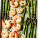 Shrimp sheet pan with asparagus - the image shows a baking tray filled with cooked shrimp and asparagus. the shrimp are pink and appear to be seasoned with herbs and spices. the tray is lined with parchment paper and there is a slice of lemon on the side. the background is a white marble countertop.