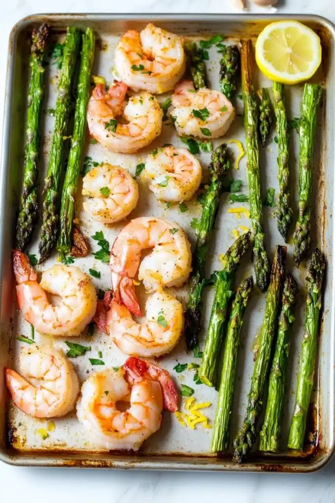 Shrimp sheet pan with asparagus - the image shows a baking tray filled with cooked shrimp and asparagus. the shrimp are pink and appear to be seasoned with herbs and spices. the tray is lined with parchment paper and there is a slice of lemon on the side. the background is a white marble countertop.