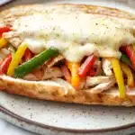 Skillet chicken cheesesteak - the image shows a close-up of a sandwich on a white plate. the sandwich appears to be made with a toasted bread and is filled with shredded chicken, red and yellow bell peppers, and green onions. the chicken is cooked to a golden brown color and is oozing out of the sides. the peppers are bright red and appear to be seasoned with herbs and spices. the cheese is melted and bubbly on top of the sandwich. the plate is sitting on a marble countertop with a blurred background.
