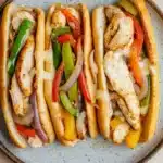 Skillet chicken cheesesteak - the image shows three hot dogs on a blue and white speckled plate. the hot dogs are filled with grilled chicken, red and green bell peppers, onions, and cheese. the chicken is cooked to a golden brown and appears to be seasoned with herbs and spices. the peppers are bright red and yellow, and the onions are sliced into thin strips. the cheese is melted and oozing out of the sides of the hot dogs. the plate is sitting on a wooden table.