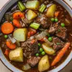 Slow cooker beef and vegetables stew - the image is a close-up of a bowl of beef stew. the stew is made with chunks of beef, potatoes, carrots, celery, and peas. the beef is cooked medium-rare and appears to be seasoned with herbs and spices. the potatoes are yellow and the carrots are orange. the peas are green and look fresh. the broth is a rich brown color and is thick and thick. the bowl is sitting on a wooden table with a sprig of parsley scattered around it.