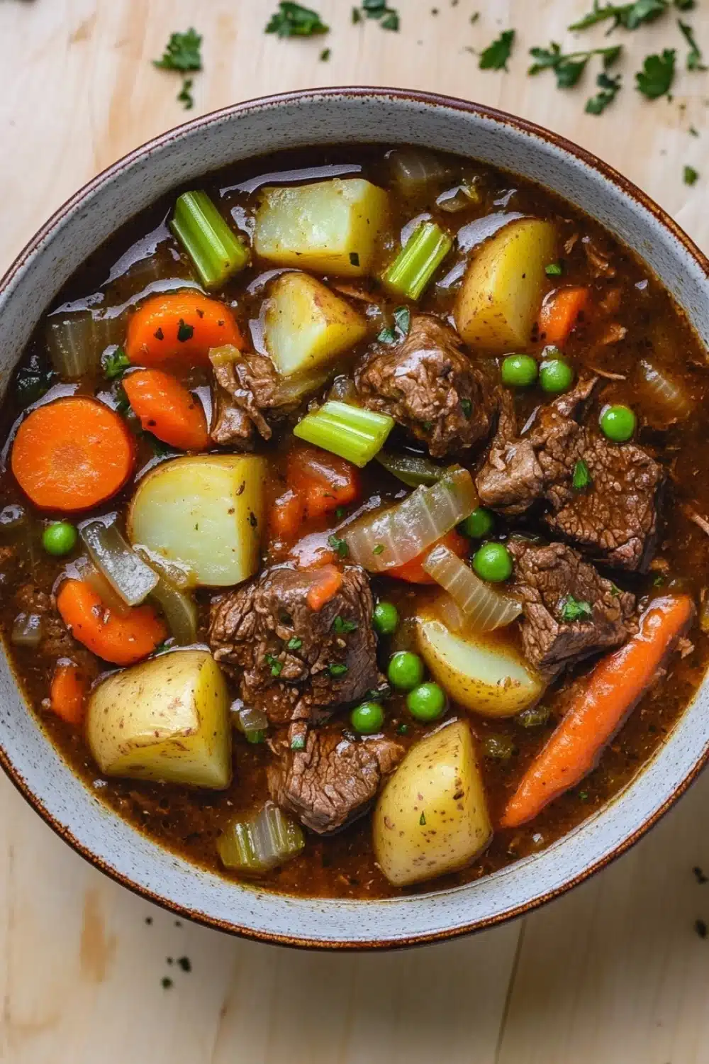 Slow cooker beef and vegetables stew - the image is a close-up of a bowl of beef stew. the stew is made with chunks of beef, potatoes, carrots, celery, and peas. the beef is cooked medium-rare and appears to be seasoned with herbs and spices. the potatoes are yellow and the carrots are orange. the peas are green and look fresh. the broth is a rich brown color and is thick and thick. the bowl is sitting on a wooden table with a sprig of parsley scattered around it.