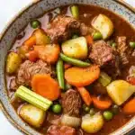 Slow cooker beef and vegetables stew - the image is a close-up of a bowl of beef stew. the stew is a rich, dark brown color and appears to be thick and hearty. it is filled with chunks of beef, carrots, potatoes, and green peas. the beef is cooked to a medium-rare and has a tender texture. the vegetables are mixed in with the stew, creating a colorful and appetizing dish. the bowl is sitting on a white marble countertop.