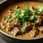 Slow cooker beef korma curry - the image is a close-up of a bowl of indian curry. the curry is a rich orange color and appears to be thick and creamy. it has chunks of meat and vegetables mixed in with a creamy sauce. the dish is garnished with fresh cilantro leaves. the bowl is made of dark-colored ceramic and is sitting on a wooden table. the background is blurred, making the curry the focal point of the image.