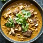 Slow cooker beef korma curry - the image is a close-up of a bowl of indian curry. the curry is a rich orange color and appears to be thick and creamy. it is garnished with fresh cilantro and sliced almonds. the bowl is made of dark blue ceramic and is sitting on a dark grey textured surface. there are a few sprigs of cilantro scattered around the bowl. the overall mood of the image is warm and inviting.