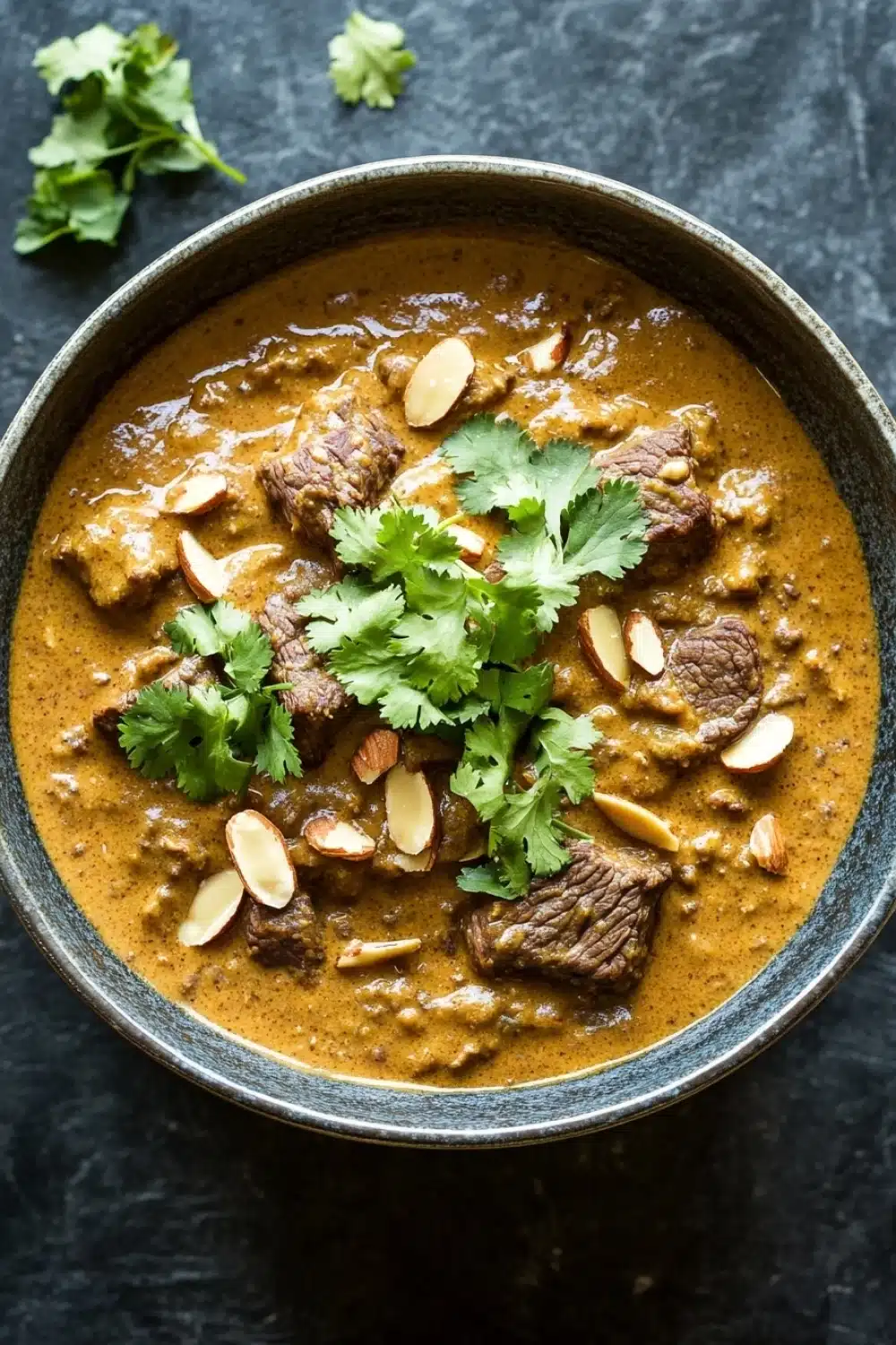 Slow cooker beef korma curry - the image is a close-up of a bowl of indian curry. the curry is a rich orange color and appears to be thick and creamy. it is garnished with fresh cilantro and sliced almonds. the bowl is made of dark blue ceramic and is sitting on a dark grey textured surface. there are a few sprigs of cilantro scattered around the bowl. the overall mood of the image is warm and inviting.