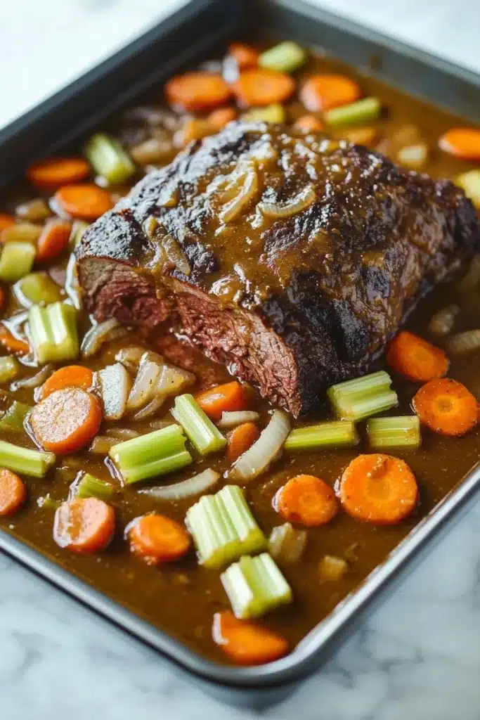 Slow cooker beef roast and gravy - the image shows a large piece of roast beef on a baking tray. the roast beef is cooked medium-rare and has a dark brown glaze on top. it is surrounded by a bed of diced carrots, celery, and onions. the carrots are bright orange and the celery is a vibrant green. the onions are a light brown color and are scattered throughout the dish. the dish is garnished with a sprinkle of herbs. the tray is sitting on a white marble countertop.