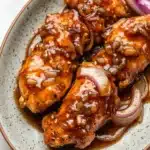Slow cooker chicken tenderloin - the image shows a plate of grilled chicken wings covered in a brown sauce. the chicken wings are arranged in a circular pattern on a white plate with a speckled rim. the sauce appears to be a deep orange color and is drizzled over the wings. on top of the chicken wings, there are sliced red onions and sliced onions. the plate is sitting on a marble countertop.