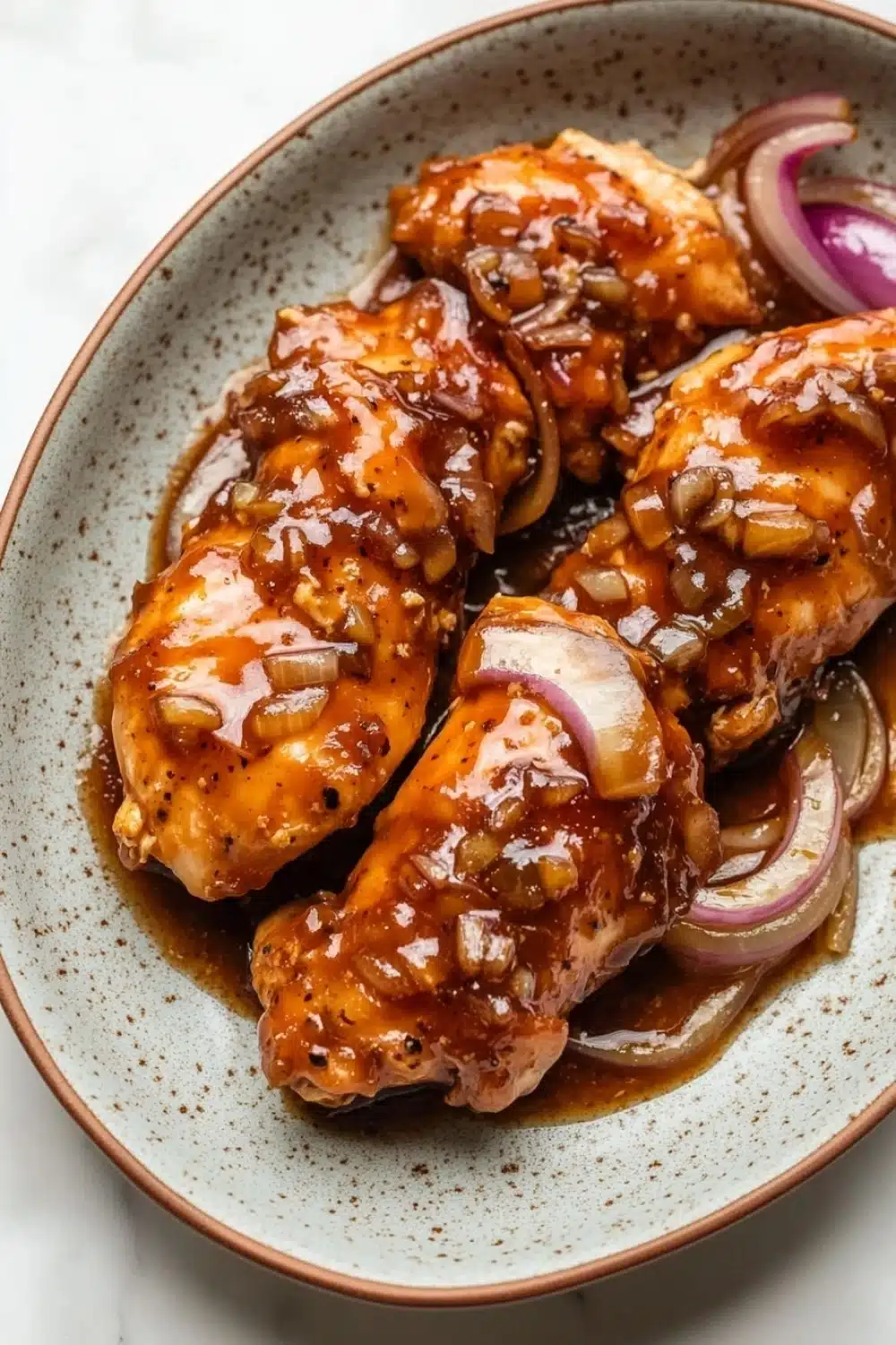 Slow cooker chicken tenderloin - the image shows a plate of grilled chicken wings covered in a brown sauce. the chicken wings are arranged in a circular pattern on a white plate with a speckled rim. the sauce appears to be a deep orange color and is drizzled over the wings. on top of the chicken wings, there are sliced red onions and sliced onions. the plate is sitting on a marble countertop.